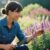 Editorial style environmental portrait, medium shot of a woman in her late 30s with a genuine smile, kneeling beside a lush, natural-looking perennial bed in her garden. She's holding a small trowel and gently touching a blooming salvia. The lighting is soft morning sun, creating a gentle bokeh effect in the background with other garden elements. The overall mood is one of quiet satisfaction and joy. True-to-life colors, shot on a 35mm camera, Kodak Portra 400 style with slight film grain. The focus is on her authentic emotion and connection to the plants, visible skin texture.