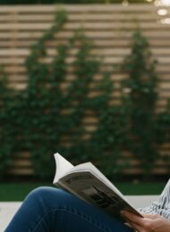 Sichtschutz Garten Ideen für eine private Wohlfühloase in einem modernen Garten. High resolution photography, Medium Shot einer entspannten Frau mittleren Alters, die auf einer stilvollen Holzterrasse ein Buch liest. Im unscharfen Hintergrund ist ein moderner Sichtschutz aus horizontalen Holzlatten zu sehen, der mit Kletterpflanzen bewachsen ist. Das warme Licht der späten Nachmittagssonne schafft eine gemütliche, intime Atmosphäre. Der Fokus liegt auf dem Gefühl der Geborgenheit und Ruhe. Editorial Style, shot on 35mm camera, Kodak Portra 400 style, muted tones, visible skin texture, depth of field.
